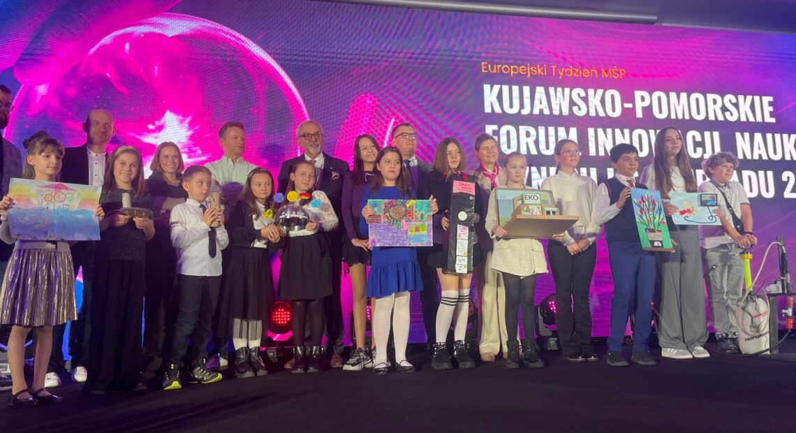 Grupowe zdjęcie na scenie podczas gali laureatów konkursu Mój Innowacyjny Wynalazek 2025. W pierwszym rzędzie stoi kilkanaście dzieci i nastolatków, każde trzyma swój projekt lub makietę wynalazku. Za nimi stoją dorośli opiekunowie i organizatorzy wydarzenia. W tle widać duży, kolorowy ekran z napisem Kujawsko-Pomorskie Forum Innowacji Nauki Biznesu i Samorządu