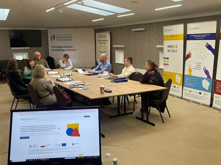 Uczestnicy szkolenia w trakcie dyskusji przy dużym, drewnianym stole. W tle widoczne są banery projektu. Na pierwszym planie na ekranie laptopa widoczna jest strona internetowa Funduszu Pożyczkowego „Po Prostu Biznes”.