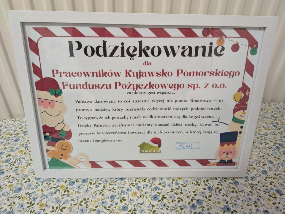Zbliżenie na oprawiony w białą ramę certyfikat z podziękowaniem od Fundacji Hospicjum Nadzieja. Tło podziękowania jest białe z czerwono-białymi pasami u góry i motywami świątecznymi po bokach. Główny tekst, czarną czcionką, brzmi: "Podziękowanie dla Pracowników Kujawsko-Pomorskiego Funduszu Pożyczkowego sp. z o.o. za piękny gest wsparcia." Poniżej znajduje się dodatkowy tekst wyjaśniający znaczenie wsparcia. Na dole widoczne jest logo Fundacji Nadzieja oraz podpis PREZES ZARZĄDU. Certyfikat leży na wzorzystym, jasnym materiale.