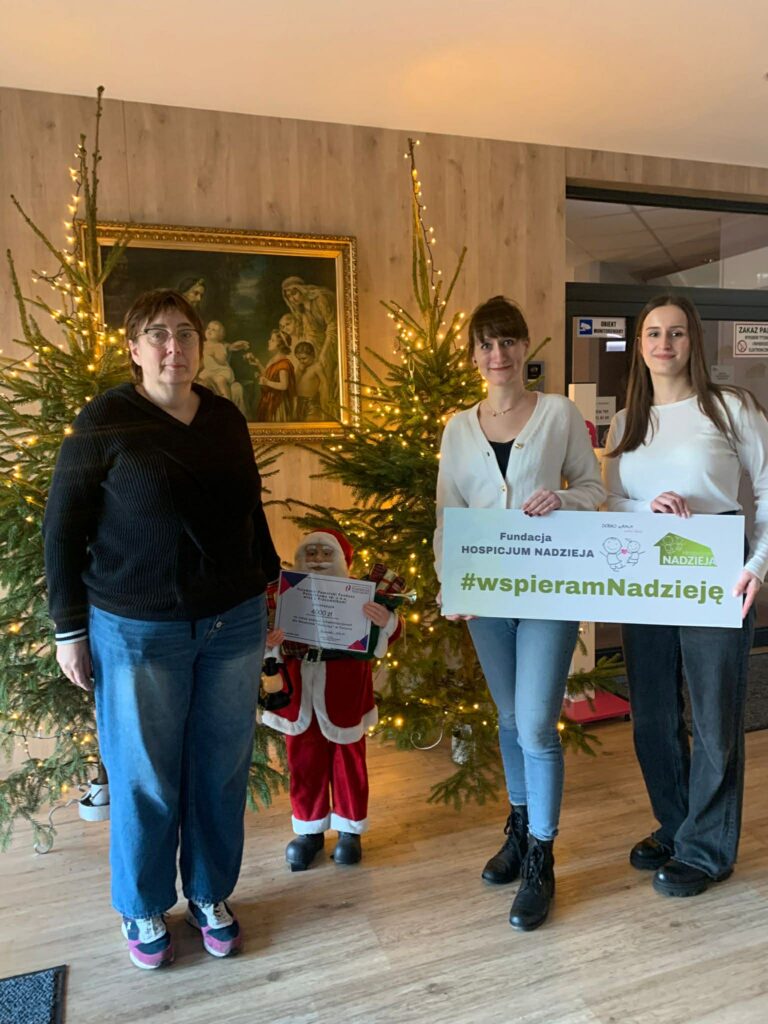 Trzy kobiety stoją wewnątrz pomieszczenia obok dwóch udekorowanych choinek i ramy z obrazem. Kobieta po lewej stronie, w ciemnym swetrze i niebieskich dżinsach, stoi przodem do obiektywu. Po prawej stronie dwie kobiety w jasnych bluzkach i dżinsach trzymają poziomy, biały transparent z logo Fundacji Hospicjum Nadzieja i napisem: "Fundacja HOSPICJUM NADZIEJA #wspieramNADZIEJĘ". Pomiędzy pierwszą kobietą a dwoma pozostałymi znajduje się mała figurka Świętego Mikołaja trzymająca w dłoni oprawiony certyfikat z podziękowaniem. Zbliżenie pokazuje, że jest to "Podziękowanie dla Pracowników Kujawsko-Pomorskiego Funduszu Pożyczkowego sp. z o.o. za piękny gest wsparcia". W tle widoczny jest obraz o tematyce religijnej w złotej ramie oraz szklane drzwi wejściowe.