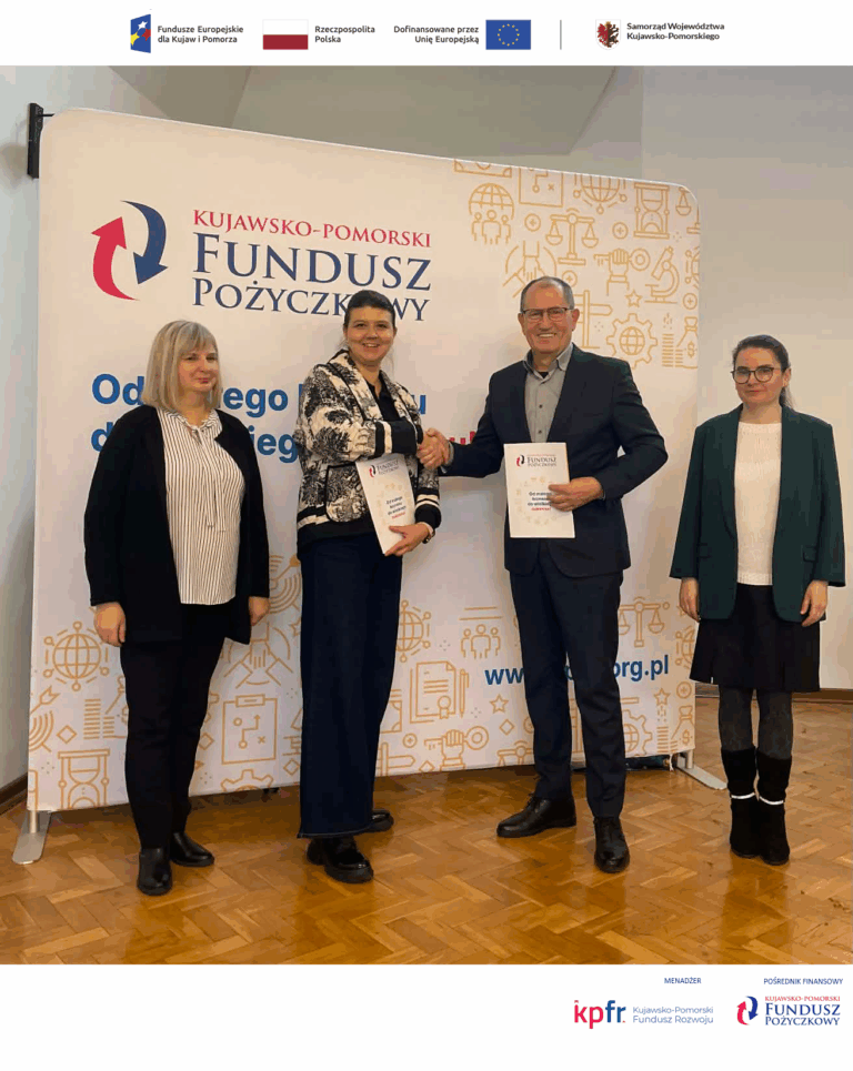 Na zdjęciu widać cztery osoby stojące na tle baneru informacyjnego Kujawsko-Pomorskiego Funduszu Pożyczkowego. Osoby w centrum uściskają sobie dłonie i trzymają dokumenty, co symbolizuje podpisanie umowy. Góra baneru zawiera logotypy: Fundusze Europejskie dla Kujaw i Pomorza, Rzeczpospolita Polska, Unia Europejska oraz Samorząd Województwa Kujawsko-Pomorskiego. Dół zdjęcia zawiera logotypy: MENADŻER (Kujawsko-Pomorski Fundusz Rozwoju) i POŚREDNIK FINANSOWY (Kujawsko-Pomorski Fundusz Pożyczkowy).