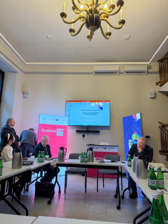 Uczestnicy spotkania siedzą przy stołach ustawionych w literę U w jasnym pomieszczeniu z ozdobnym żyrandolem. Na środkowej ścianie wisi telewizor z wyświetloną prezentacją o polityce energetycznej województwa. Po obu stronach ekranu stoją wysokie banery reklamowe: różowy z napisem „Business in Kujawy & Pomorze” oraz niebieski z grafiką geometryczną. Na stołach znajdują się termosy, filiżanki i butelki wody mineralnej.