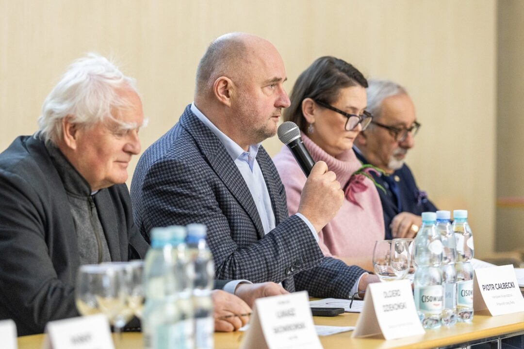 Piotr Całbecki, Marszałek Województwa Kujawsko-Pomorskiego, przemawia do mikrofonu podczas oficjalnego spotkania. Obok niego przy stole siedzą inni uczestnicy panelu, w tym Olgierd Dziekoński, przed którymi ustawiono tabliczki z nazwiskami i wodę mineralną.