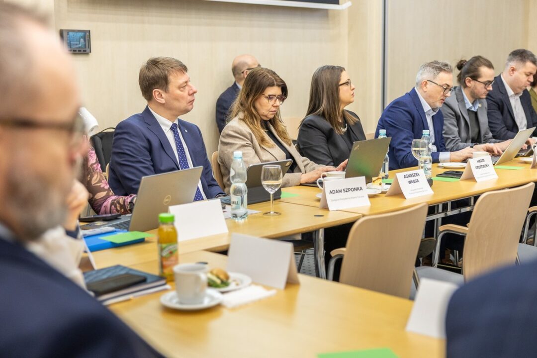 Uczestnicy spotkania, w tym Adam Szponka, Agnieszka Wasita i Marta Krużewska, siedzą przy długim stole z laptopami i notatkami. Skupieni słuchają wystąpienia podczas oficjalnej narady w sali konferencyjnej.