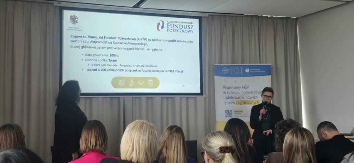 Sala konferencyjna wypełniona ludźmi. Na ekranie slajd Kujawsko-Pomorskiego Funduszu Pożyczkowego z informacjami o wspieraniu biznesu w regionie i udzieleniu ponad 4700 pożyczek. Obok ekranu stoi kobieta z mikrofonem i baner z napisem: Wspieramy MŚP w rozwoju innowacyjności i zdobywaniu nowych rynków zagranicznych.