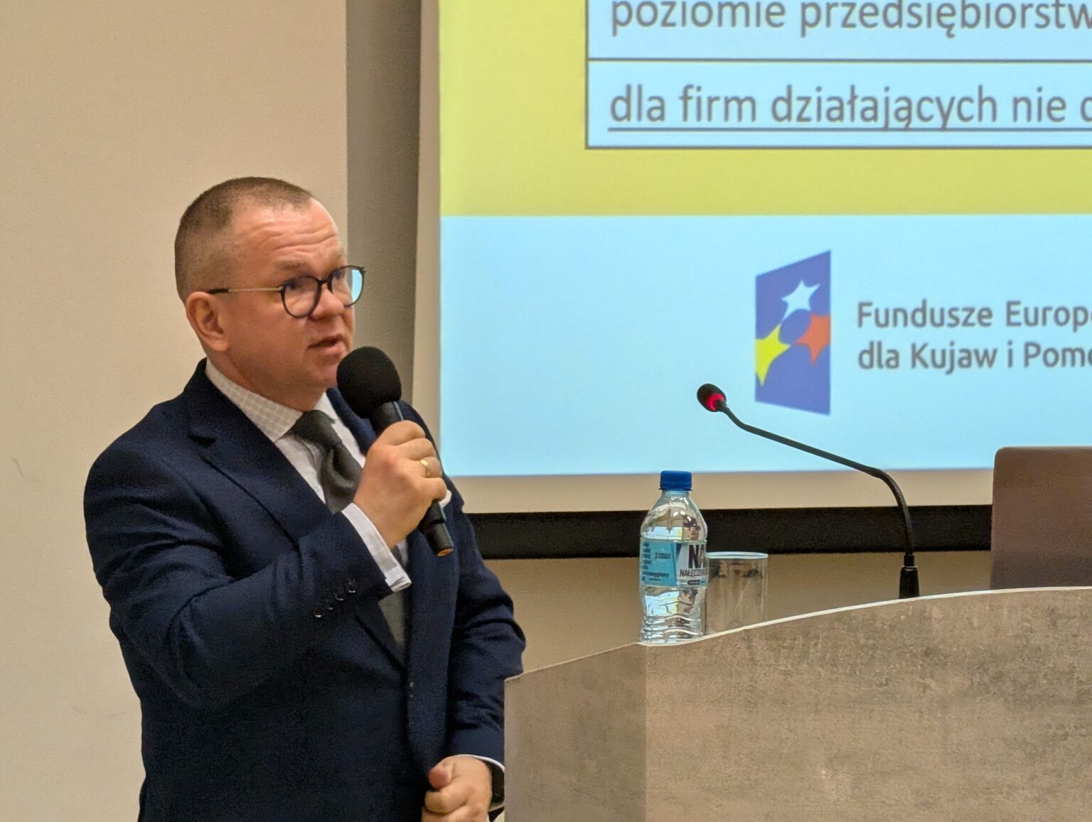 Zbliżenie na mężczyznę w okularach przemawiającego do mikrofonu przy mównicy. W tle fragment prezentacji z logo Funduszy Europejskich dla Kujaw i Pomorza.