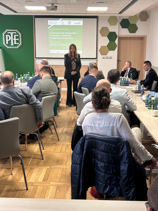 Uczestnicy spotkania siedzą przy stołach ustawionych w podkowę. Na środku, przed ekranem, stoi kobieta w ciemnym garniturze. Na ekranie napis: Fundusze EU dla rozwoju MŚP – Innowacje, rozwój kadr, inteligentne specjalizacje. Na bocznej ścianie logo PTE oraz dekoracje w kształcie heksagonów.