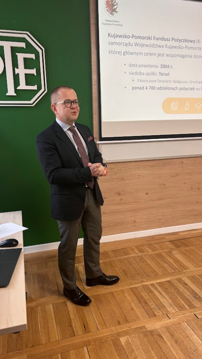 Mężczyzna w okularach, ciemnej marynarce i krawacie przemawia, stojąc obok ekranu projekcyjnego. Na ekranie informacje o Kujawsko-Pomorskim Funduszu Pożyczkowym, m.in. data powstania 2004 r. i siedziba w Toruniu. Po lewej stronie na zielonej ścianie widoczne białe logo PTE.