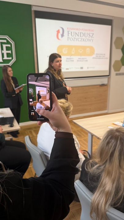 Osoba na pierwszym planie trzyma smartfon i robi zdjęcie kobiecie przemawiającej podczas spotkania w sali konferencyjnej. W tle widać ekran projekcyjny z logo Kujawsko-Pomorskiego Funduszu Pożyczkowego oraz napisami: Granty dla firm i Projekty edukacyjne. Po lewej stronie, na zielonej ścianie, widnieje fragment białego logo PTE.