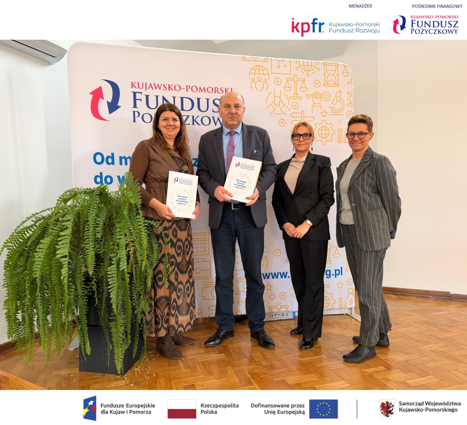 Cztery osoby – trzy kobiety i mężczyzna – stoją na tle ścianki reklamowej Kujawsko-Pomorskiego Funduszu Pożyczkowego. Mężczyzna i jedna z kobiet trzymają białe teczki z logo funduszu i napisem „Od małego biznesu do wielkiego sukcesu!”. Na dole logotypy informujące o dofinansowaniu pożyczki ze środków Unii Europejskiej i Samorządu Województwa Kujawsko-Pomorskiego. U góry logotypy Menadżera: KPFR Kujawsko-Pomorski Fundusz Rozwoju, i pośrednika finansowego: Kujawsko-Pomorskiego Funduszu Pożyczkowego.