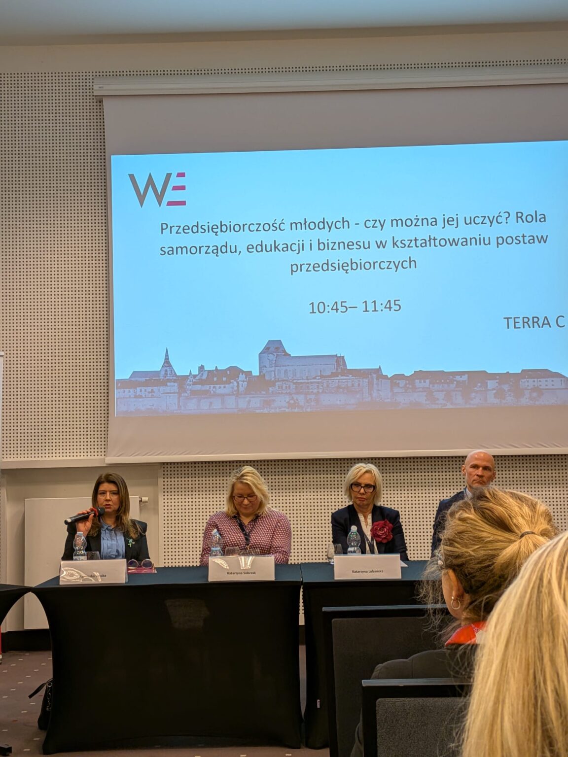 Panel dyskusyjny podczas konferencji. Cztery osoby siedzą za stołem z tabliczkami imiennymi: Agnieszka Wrońska, Katarzyna Sobczak i Katarzyna Lubańska. Pierwsza z lewej kobieta mówi do mikrofonu. W tle ekran z napisem: Przedsiębiorczość młodych – czy można jej uczyć? Rola samorządu, edukacji i biznesu w kształtowaniu postaw przedsiębiorczych.