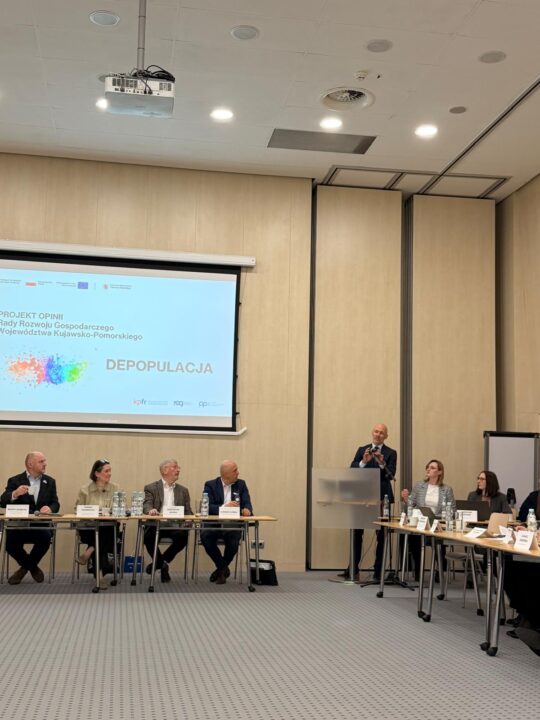 Szerokie ujęcie sali konferencyjnej, w której na ekranie projekcyjnym wyświetlana jest prezentacja pt. "PROJEKT OPINII Rady Rozwoju Gospodarczego Województwa Kujawsko-Pomorskiego", a obok na podium stoi prelegent w garniturze. Poniżej siedzą uczestnicy debaty przy stołach ułożonych w kształt litery U, a sufit jest oświetlony panelami LED.