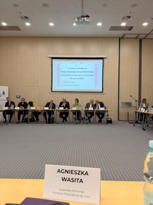 Widok z pierwszego planu na imienną tabliczkę na stole konferencyjnym z napisem "AGNIESZKA WASITA" oraz "Kujawsko-Pomorski Fundusz Pożyczkowy sp. z o.o.". W tle widać rzędy siedzących uczestników posiedzenia oraz ekran z prezentacją z datą "25 marca 2026 r.", w przestronnej, jasno oświetlonej sali.