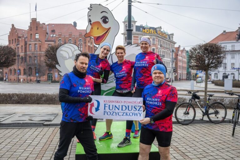 Grupa pięciorga biegaczy w niebiesko-czerwonych koszulkach z napisem „Kujawy Pomorze” pozuje przy planszy z uśmiechniętą maskotką gęsi. Z przodu trzymają baner Kujawsko-Pomorskiego Funduszu Pożyczkowego na tle architektury Torunia.