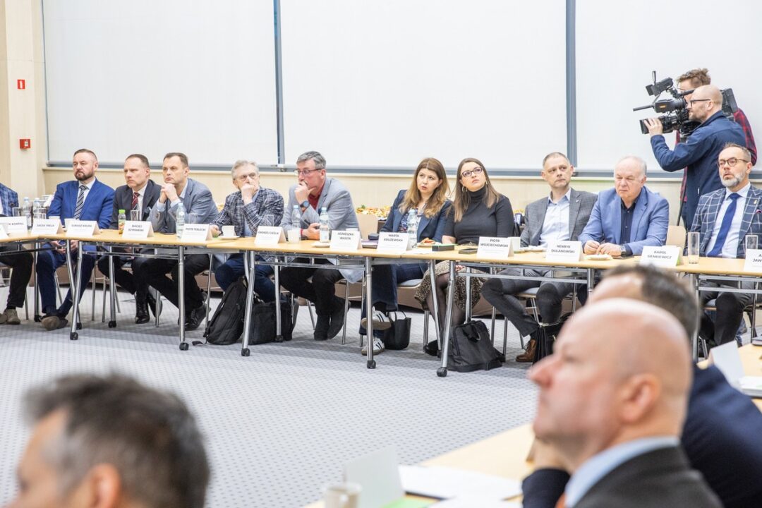 Zdjęcie grupy ekspertów i urzędników siedzących wzdłuż stołów konferencyjnych podczas oficjalnego posiedzenia w nowoczesnej sali. Uczestnicy są ubrani w stroje biznesowe i dyskutują, a przed nimi widoczne są tabliczki z nazwiskami, woda butelkowana i sprzęt nagrywający, w tym kamera w tle.