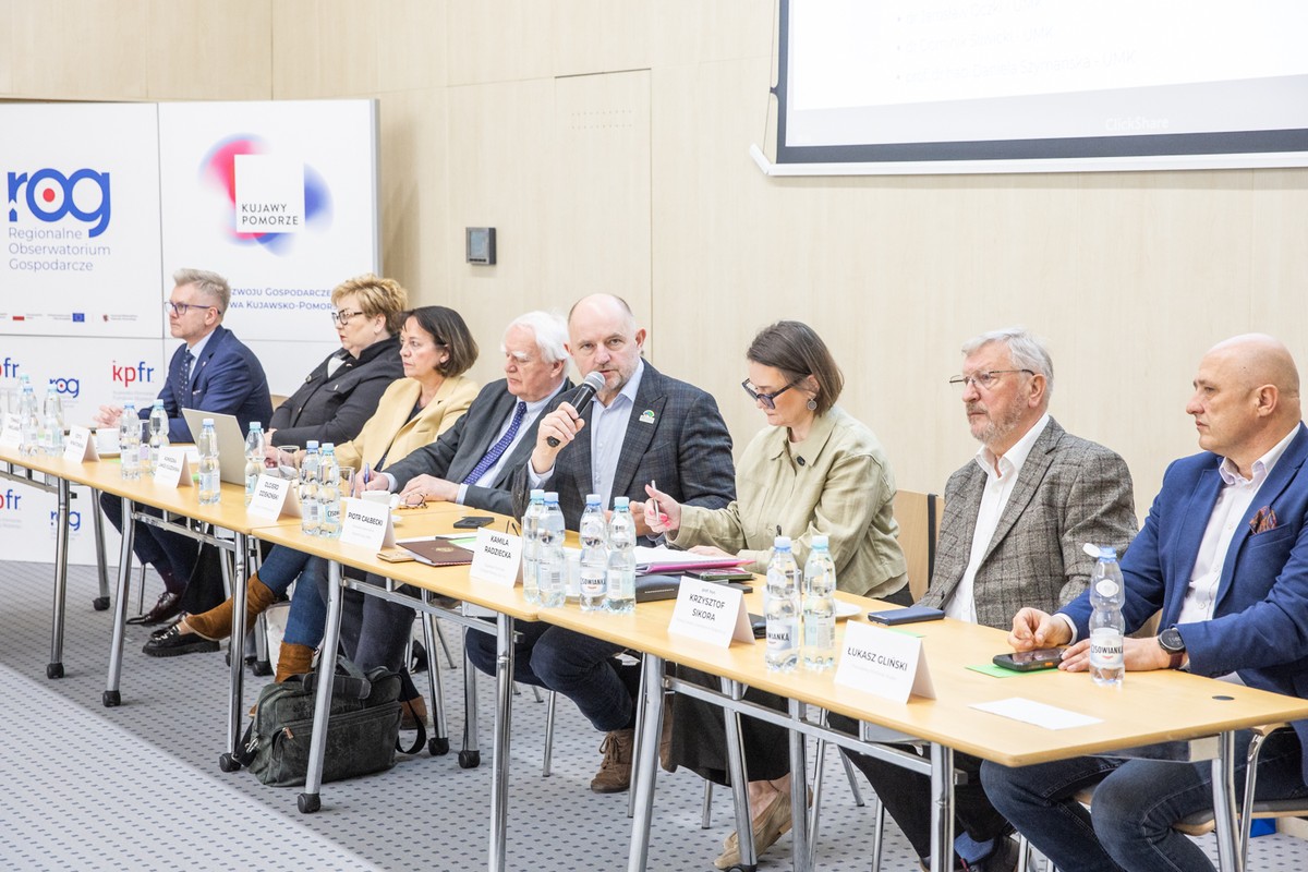 Zbliżenie na prelegenta w kraciastej marynarce i dżinsach, przemawiającego do mikrofonu przy stole konferencyjnym. Obok niego siedzą inni uczestnicy spotkania, a w tle widać ekran z logiem "Kujawy Pomorze" oraz butelki wody mineralnej "Cisowianka" na pierwszym planie.