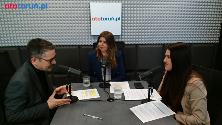 Trzy osoby siedzące przy stole w studiu nagraniowym z logo ototorun.pl na ścianie.