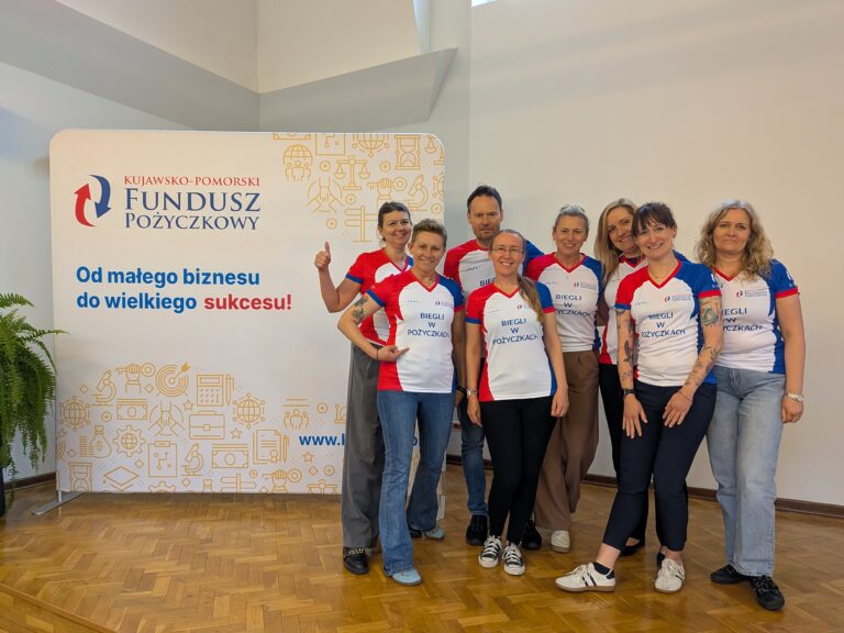 Grupa ośmiu uśmiechniętych osób pozuje do wspólnego zdjęcia w pomieszczeniu z drewnianym parkietem, stojąc obok białej ścianki reklamowej Kujawsko-Pomorskiego Funduszu Pożyczkowego z hasłem „Od małego biznesu do wielkiego sukcesu!”. Większość osób ma na sobie jednakowe, biało-czerwono-niebieskie sportowe koszulki z napisem „BIEGLI W POŻYCZKACH” i logo instytucji.