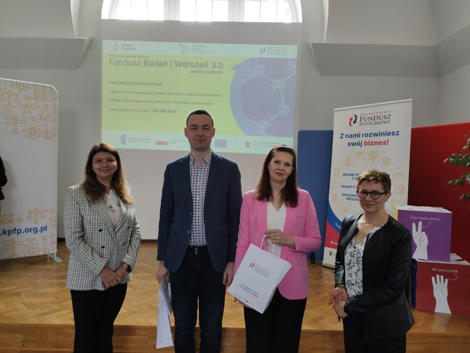 Zdjęcie z uroczystości podpisania umów grantowych w ramach projektu Fundusz Badań i Wdrożeń 3.0. Na podium stoją przedstawiciele Kujawsko-Pomorskiego Funduszu Pożyczkowego (grantodawcy) oraz beneficjenci, pozując na tle ekranu z informacjami o przyznanym dofinansowaniu i banerów promujących Kujawsko-Pomorski Fundusz Pożyczkowy. Uczestnicy trzymają teczki z dokumentami lub torebki upominkowe.