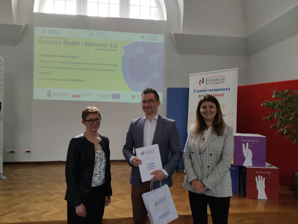 Zdjęcie z uroczystości podpisania umów grantowych w ramach projektu Fundusz Badań i Wdrożeń 3.0. Na podium stoją przedstawiciele Kujawsko-Pomorskiego Funduszu Pożyczkowego (grantodawcy) oraz beneficjenci, pozując na tle ekranu z informacjami o przyznanym dofinansowaniu i banerów promujących Kujawsko-Pomorski Fundusz Pożyczkowy. Uczestnicy trzymają teczki z dokumentami lub torebki upominkowe.