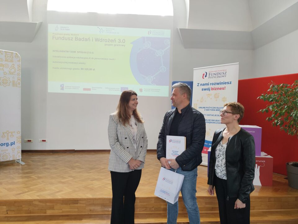 Zdjęcie z uroczystości podpisania umów grantowych w ramach projektu Fundusz Badań i Wdrożeń 3.0. Na podium stoją przedstawiciele Kujawsko-Pomorskiego Funduszu Pożyczkowego (grantodawcy) oraz beneficjenci, pozując na tle ekranu z informacjami o przyznanym dofinansowaniu i banerów promujących Kujawsko-Pomorski Fundusz Pożyczkowy. Uczestnicy trzymają teczki z dokumentami lub torebki upominkowe.