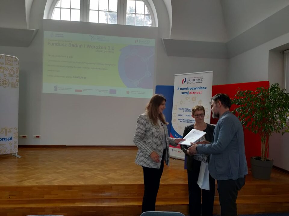 Zdjęcie z uroczystości podpisania umów grantowych w ramach projektu Fundusz Badań i Wdrożeń 3.0. Na podium stoją przedstawiciele Kujawsko-Pomorskiego Funduszu Pożyczkowego (grantodawcy) oraz beneficjenci, pozując na tle ekranu z informacjami o przyznanym dofinansowaniu i banerów promujących Kujawsko-Pomorski Fundusz Pożyczkowy. Uczestnicy trzymają teczki z dokumentami lub torebki upominkowe.