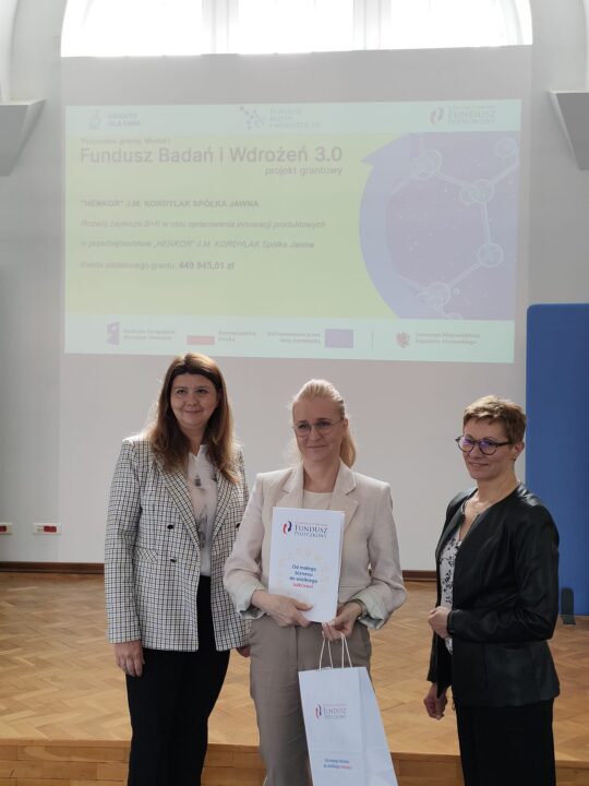 Zdjęcie z uroczystości podpisania umów grantowych w ramach projektu Fundusz Badań i Wdrożeń 3.0. Na podium stoją przedstawiciele Kujawsko-Pomorskiego Funduszu Pożyczkowego (grantodawcy) oraz beneficjenci, pozując na tle ekranu z informacjami o przyznanym dofinansowaniu i banerów promujących Kujawsko-Pomorski Fundusz Pożyczkowy. Uczestnicy trzymają teczki z dokumentami lub torebki upominkowe.