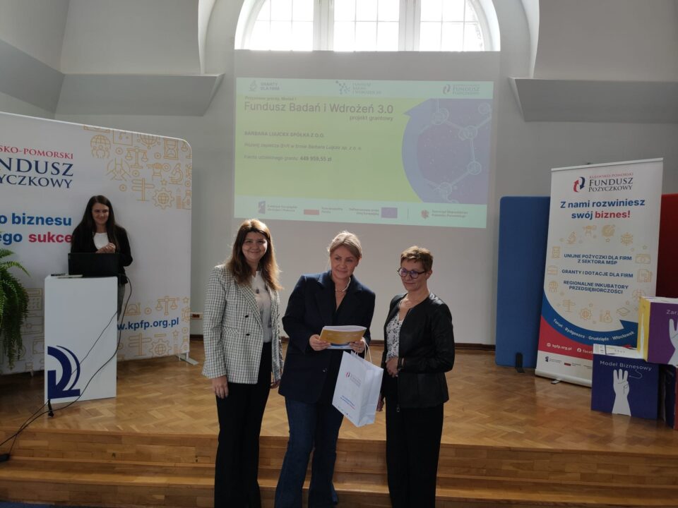 Zdjęcie z uroczystości podpisania umów grantowych w ramach projektu Fundusz Badań i Wdrożeń 3.0. Na podium stoją przedstawiciele Kujawsko-Pomorskiego Funduszu Pożyczkowego (grantodawcy) oraz beneficjenci, pozując na tle ekranu z informacjami o przyznanym dofinansowaniu i banerów promujących Kujawsko-Pomorski Fundusz Pożyczkowy. Uczestnicy trzymają teczki z dokumentami lub torebki upominkowe.