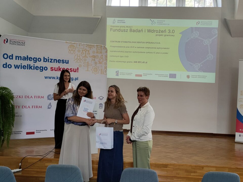 Zdjęcie z uroczystości podpisania umów grantowych w ramach projektu Fundusz Badań i Wdrożeń 3.0. Na podium stoją przedstawiciele Kujawsko-Pomorskiego Funduszu Pożyczkowego (grantodawcy) oraz beneficjenci, pozując na tle ekranu z informacjami o przyznanym dofinansowaniu i banerów promujących Kujawsko-Pomorski Fundusz Pożyczkowy. Uczestnicy trzymają teczki z dokumentami lub torebki upominkowe.