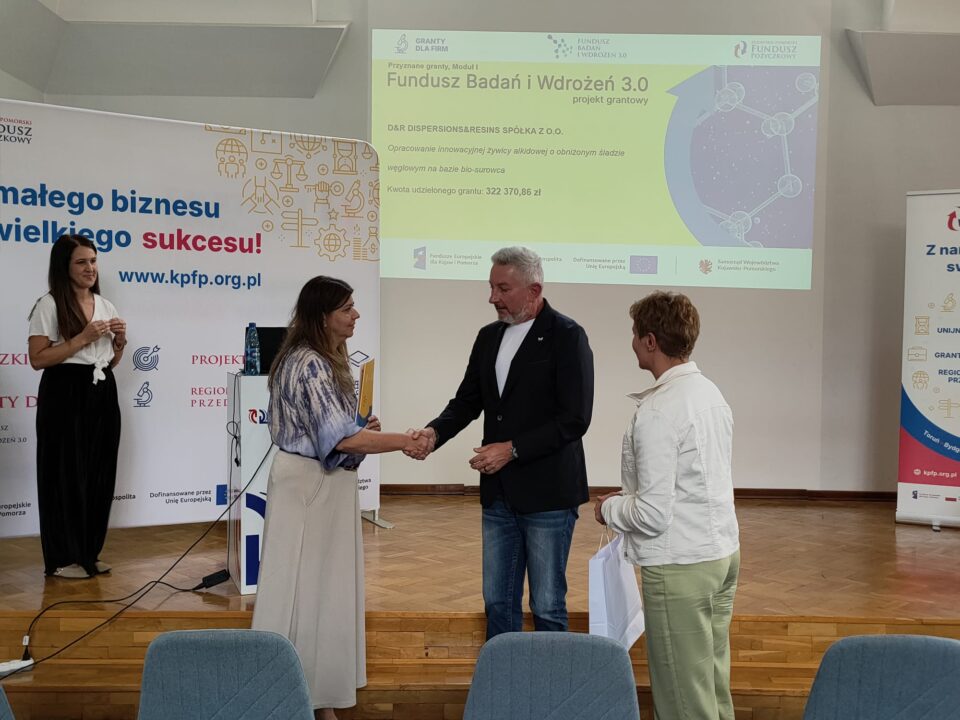 Zdjęcie z uroczystości podpisania umów grantowych w ramach projektu Fundusz Badań i Wdrożeń 3.0. Na podium stoją przedstawiciele Kujawsko-Pomorskiego Funduszu Pożyczkowego (grantodawcy) oraz beneficjenci, pozując na tle ekranu z informacjami o przyznanym dofinansowaniu i banerów promujących Kujawsko-Pomorski Fundusz Pożyczkowy. Uczestnicy trzymają teczki z dokumentami lub torebki upominkowe.