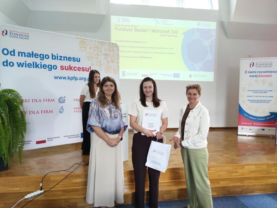 Zdjęcie z uroczystości podpisania umów grantowych w ramach projektu Fundusz Badań i Wdrożeń 3.0. Na podium stoją przedstawiciele Kujawsko-Pomorskiego Funduszu Pożyczkowego (grantodawcy) oraz beneficjenci, pozując na tle ekranu z informacjami o przyznanym dofinansowaniu i banerów promujących Kujawsko-Pomorski Fundusz Pożyczkowy. Uczestnicy trzymają teczki z dokumentami lub torebki upominkowe.
