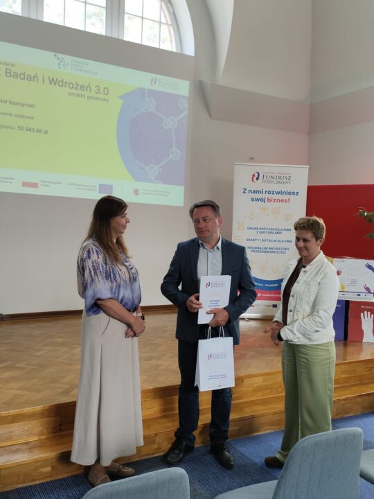 Zdjęcie z uroczystości podpisania umów grantowych w ramach projektu Fundusz Badań i Wdrożeń 3.0. Na podium stoją przedstawiciele Kujawsko-Pomorskiego Funduszu Pożyczkowego (grantodawcy) oraz beneficjenci, pozując na tle ekranu z informacjami o przyznanym dofinansowaniu i banerów promujących Kujawsko-Pomorski Fundusz Pożyczkowy. Uczestnicy trzymają teczki z dokumentami lub torebki upominkowe.