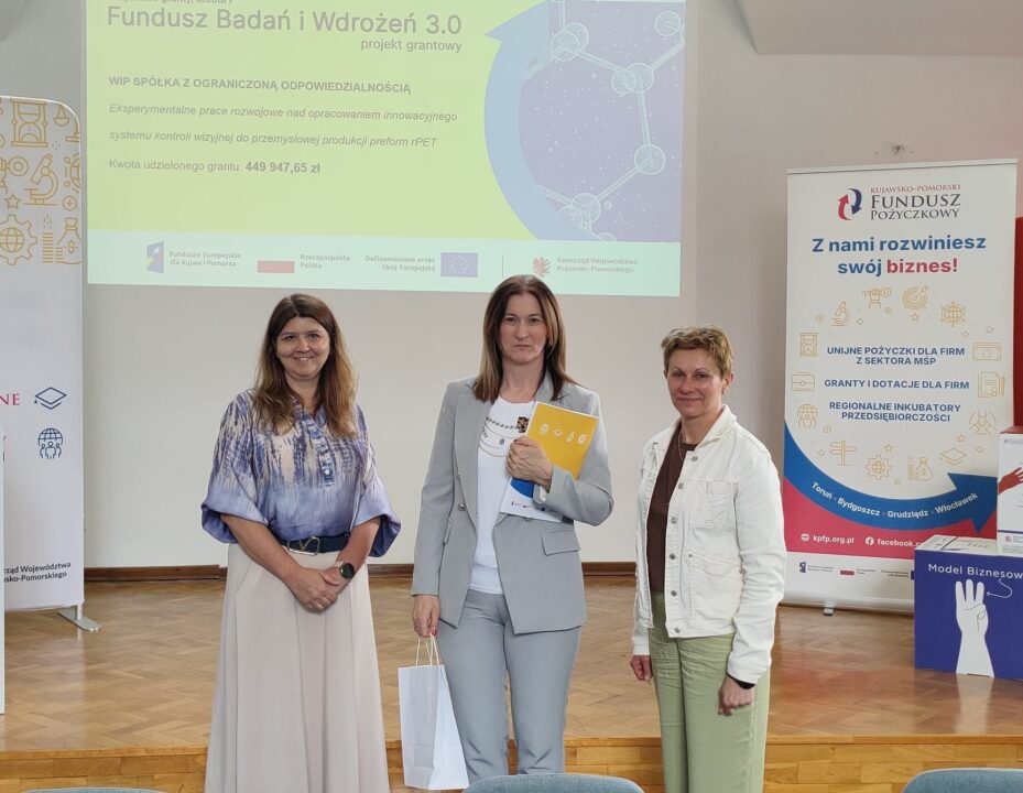 Zdjęcie z uroczystości podpisania umów grantowych w ramach projektu Fundusz Badań i Wdrożeń 3.0. Na podium stoją przedstawiciele Kujawsko-Pomorskiego Funduszu Pożyczkowego (grantodawcy) oraz beneficjenci, pozując na tle ekranu z informacjami o przyznanym dofinansowaniu i banerów promujących Kujawsko-Pomorski Fundusz Pożyczkowy. Uczestnicy trzymają teczki z dokumentami lub torebki upominkowe.