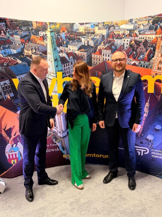 Dwie osoby, kobieta i mężczyzna, podają sobie ręce na tle wielkoformatowej grafiki przedstawiającej panoramę Torunia. Kobieta ubrana jest w ciemną koszulę i jaskrawozielone spodnie, a mężczyzna w ciemny garnitur i białą koszulę. Obok nich, po prawej stronie, stoi trzecia osoba – mężczyzna w okularach i ciemnym garniturze, patrzący w stronę obiektywu. W tle widoczne są dachy toruńskiej starówki, wieża kościoła, żółty napis oraz logotypy, w tym herb Torunia i znak Fundacji Przedsiębiorczy Toruń.