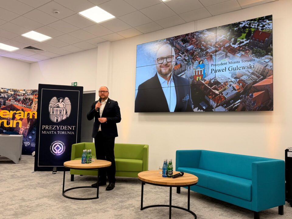 Prezydent Miasta Torunia Paweł Gulewski stoi w sali konferencyjnej i przemawia do mikrofonu. Jest ubrany w ciemny garnitur i białą koszulę. Za jego plecami znajduje się duży ekran ścienny wyświetlający grafikę z jego wizerunkiem, herbem Torunia oraz napisem „Prezydent Miasta Torunia Paweł Gulewski”. Obok prezydenta stoi pionowy baner z herbem Torunia i napisem „Prezydent Miasta Torunia”. W pomieszczeniu znajdują się dwie nowoczesne kanapy – zielona i niebieska – oraz dwa okrągłe stoliki kawowe, na których stoją butelki z wodą. W tle po lewej stronie widoczny jest fragment innej grafiki z napisem „Toruń”.