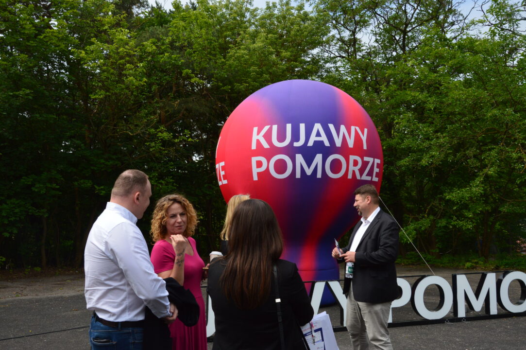 Dzień otwarty w KPFP - grupa ludzi rozmawia na tle balonu Kujawy Pomorze w ogrodzie