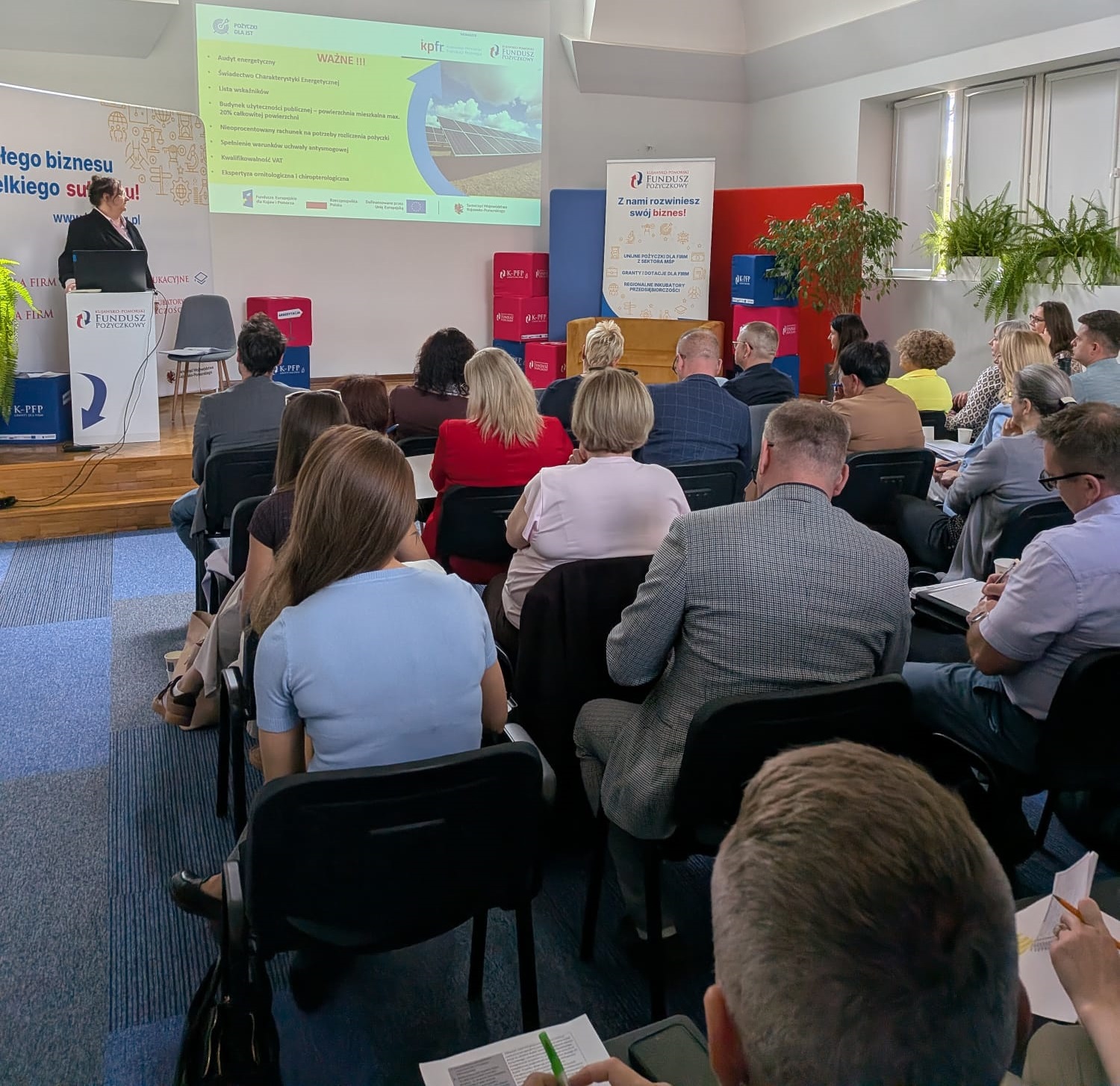 Kobieta w ciemnym żakiecie stoi przy mównicy i prowadzi prezentację podczas konferencji w jasnej sali. W tle widać duży ekran z wyświetlanym slajdem o audycie energetycznym oraz banery i pufy z logo Kujawsko-Pomorskiego Funduszu Pożyczkowego. Przed prelegentką siedzą uczestnicy spotkania widziani od tyłu, skupieni na wystąpieniu i robiący notatki.