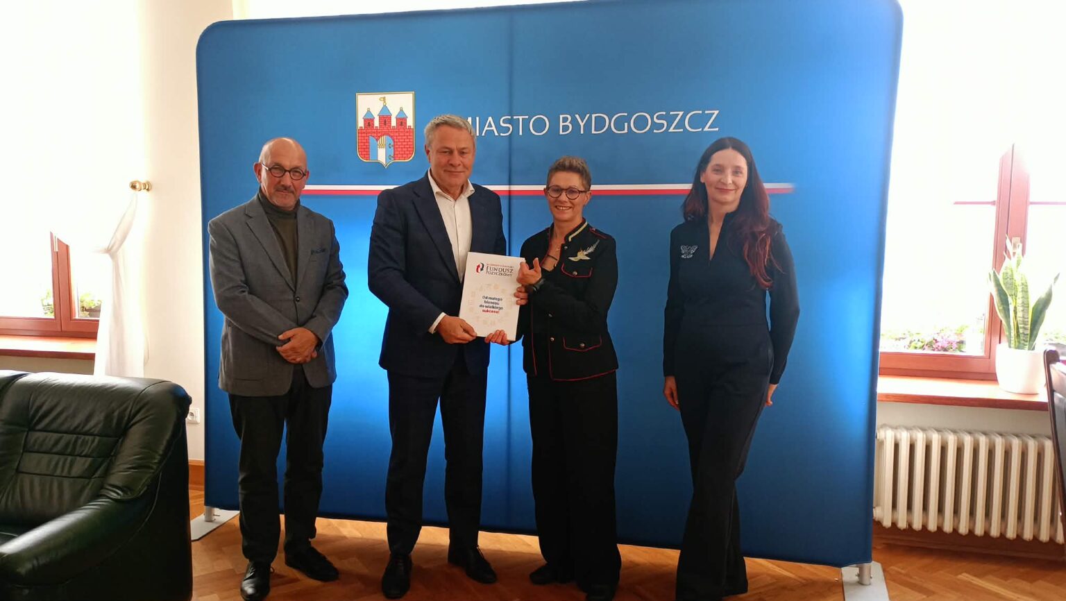 Cztery elegancko ubrane osoby (dwóch mężczyzn, dwie kobiety) pozują na tle ścianki reklamowej z napisem "Miasto Bydgoszcz". Dwie osoby trzymają teczkę z dokumentami.